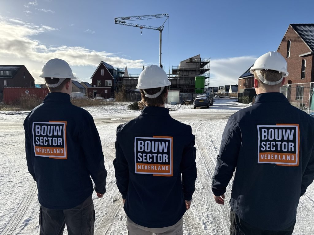 Drie vakmensen van Bouwsector Nederland in donkerblauwe werkjassen kijken uit over een besneeuwde bouwplaats